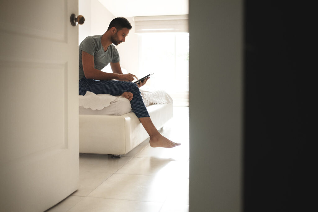 Man in casual clothing sitting on bed, looking at smartphone indoors.