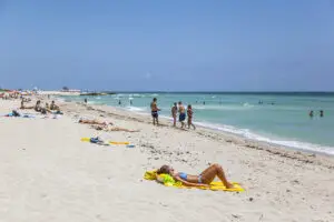 People sunbathing and swimming at a sandy beach with turquoise water under a clear blue sky.