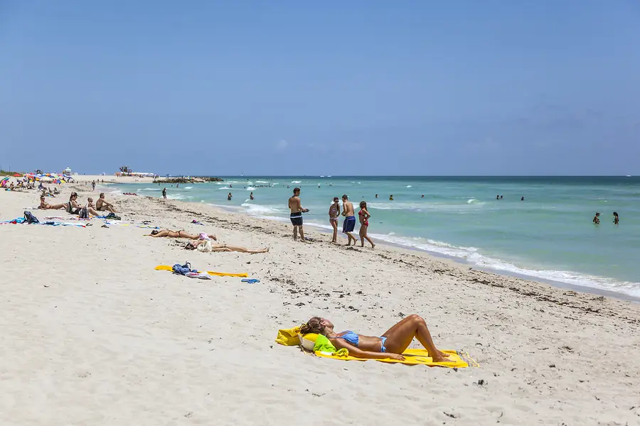 People sunbathing and swimming at a sandy beach with turquoise water under a clear blue sky.