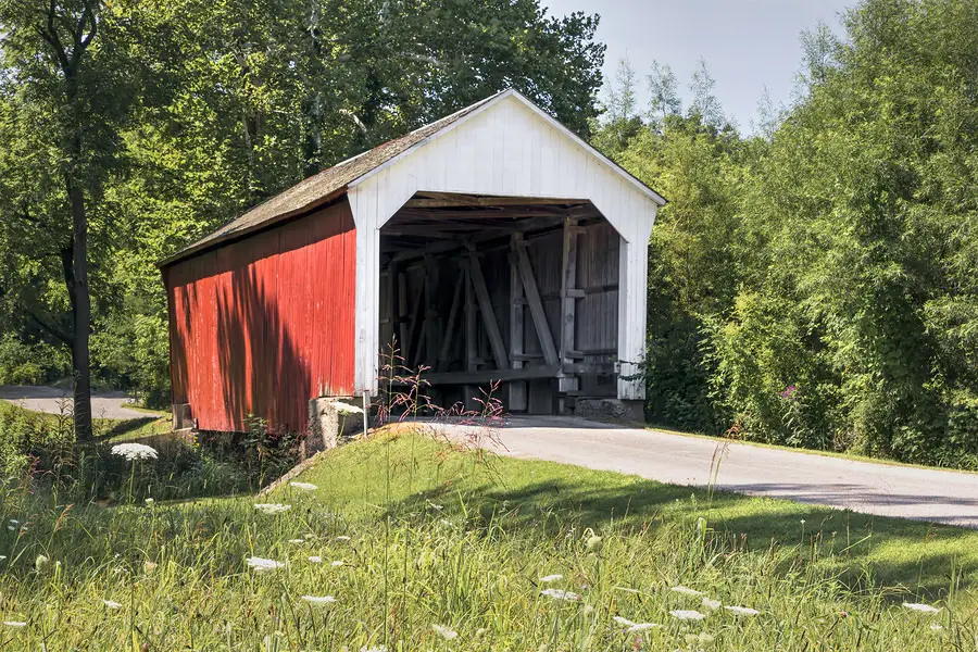 Red and white wooden covered bridge over a country road surrounded by green trees and grass.
