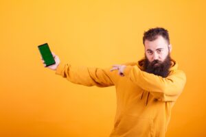 Bearded man in yellow hoodie pointing at a smartphone with a green screen against an orange background.