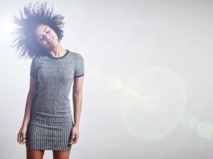 Smiling woman with curly hair tossing her head, wearing a gray striped dress against a bright white background.