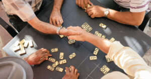 Two men shake hands over a table of dominoes while others play, symbolizing trust and personal connection.