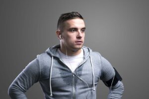 Young man in gray hoodie with earbuds and a phone armband, looking to the side against a gray background.
