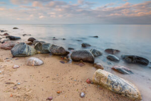 Smooth rocks and pebbles on a sandy beach meeting calm water under a soft pastel sky at dawn.