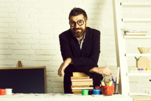 Smiling bearded man in glasses and dark blazer leaning on a stack of books at a desk against a white brick wall.