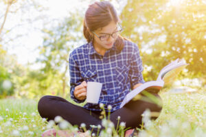 Woman in plaid shirt sitting outdoors on grass, reading a book and holding a coffee mug in sunlight.